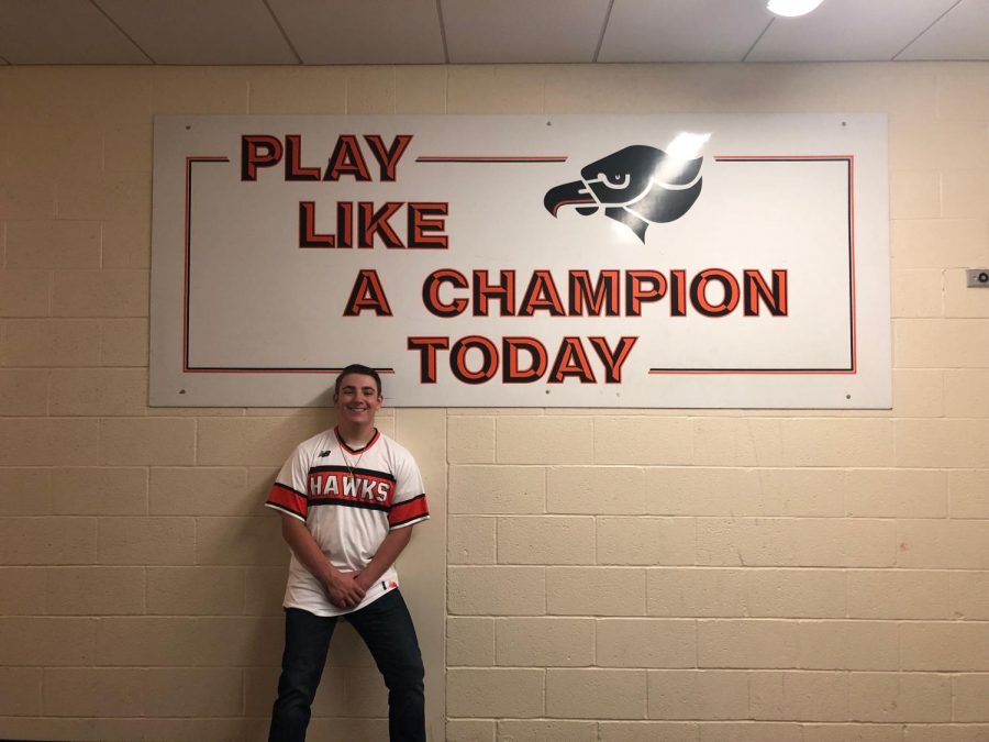 Joe Sager smiles for the camera in front of the play like a champion sign near the gym, and he is wearing his Bethel Park baseball T-shirt.